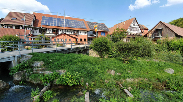 Eine malerische Ansicht eines Gebäudekomplexes mit Solarpanelen auf dem Dach, umgeben von grüner Vegetation und einem kleinen Fluss. Eine Brücke führt über den Fluss, während im Hintergrund weitere traditionelle Gebäude zu sehen sind. Der Himmel ist blau mit einigen Wolken. (Text KI generiert)