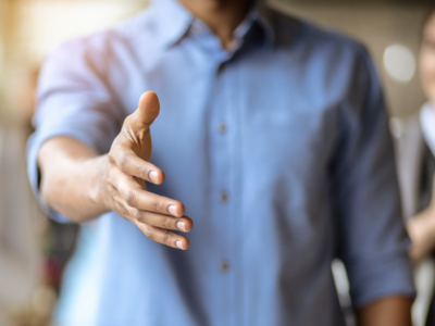 Ein Mann in einem hellblauen Hemd streckt seine Hand zum Handschlag aus. Der Hintergrund ist unscharf, wobei eine zweite Person sichtbar ist. Die Geste signalisiert eine freundliche Begrüßung oder ein Angebot zur Zusammenarbeit.