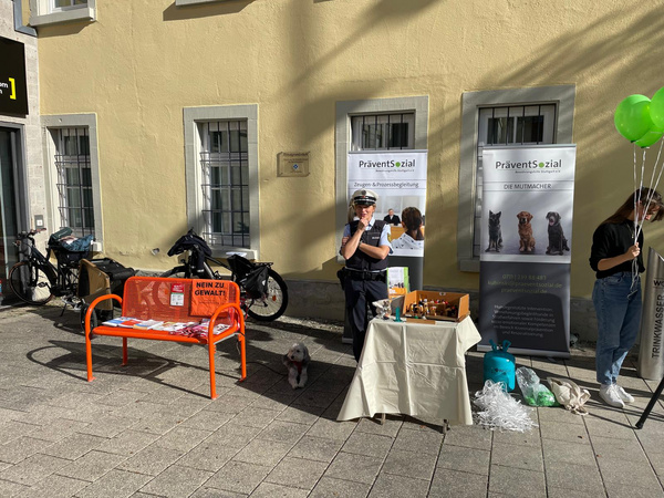 Un'agenzia di sensibilizzazione sociale è presente all'aperto, con un tavolo allestito e volantini informativi. Una poliziotta interagisce con il pubblico e un cane è seduto nei pressi. In secondo piano, si notano biciclette e palloncini verdi.