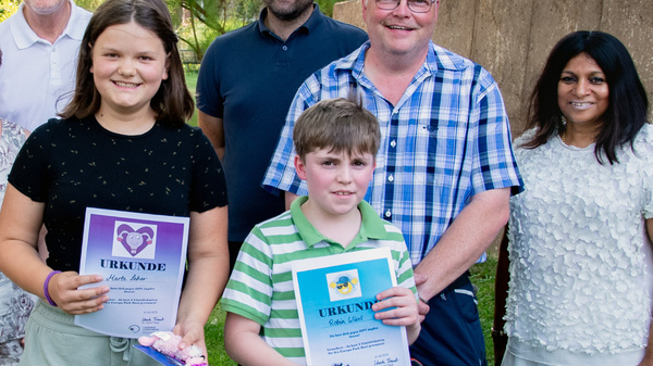 A group of eight individuals stands outdoors, smiling. Two children in the front hold certificates. The background features greenery and a brick building. The atmosphere appears joyful, suggesting a celebration or achievement recognition event.