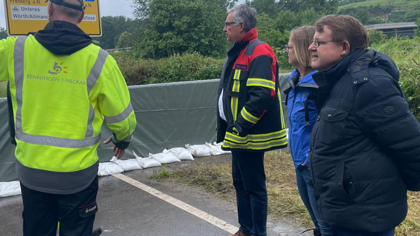 Vier Personen stehen an einer abgesperrten Straße und sprechen miteinander. Eine Person in Warnjacke zeigt auf ein Schild. Die Umgebung ist bewaldet, der Himmel ist bewölkt und es scheint, als ob es regnet.