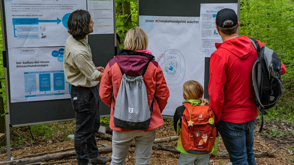 Eine Gruppe von vier Personen steht vor Informationsständen im Wald. Ein Mann erklärt das Thema Klimaschutz, während ein Plakat mit dem Titel „Unser #ClimateHandprint wächst“ im Vordergrund zu sehen ist. Die Gruppe interessiert sich sichtbar für die dargestellten Informationen.