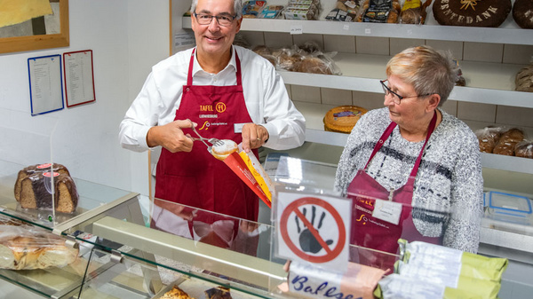 Ein freundlich lächelnder Mann und eine Frau stehen hinter einer Theke, die mit verschiedenen Broten und Backwaren gefüllt ist. Der Mann hält eine Verpackung in der Hand, während die Frau lächelnd zuschaut. Beide tragen blaue und rote Schürzen mit Aufschrift.