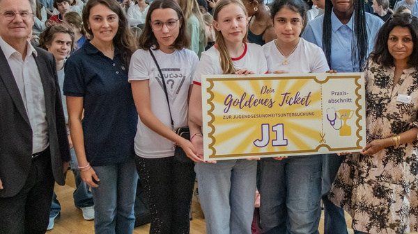 Die Gruppe von Schülern und Lehrern steht auf einer Bühne und hält ein großes Schild mit der Aufschrift "Das goldene Ticket J1". Im Hintergrund ist ein Publikum von Schülern zu sehen, das die Veranstaltung aufmerksam verfolgt.