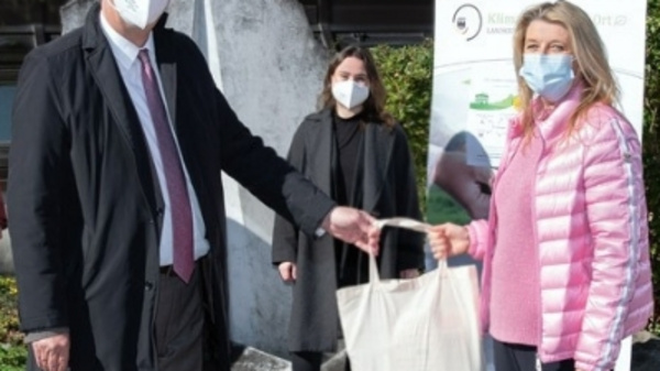 A man in a suit and mask hands a bag to a woman in a pink jacket, also wearing a mask. Behind them are two women in casual attire, all standing outdoors. The background features greenery and a sign related to health services.