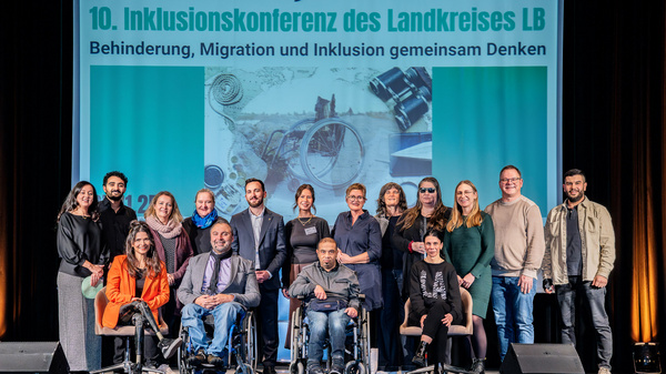Eine Gruppe von Menschen steht auf der Bühne vor einem Banner mit der Aufschrift "10. Inklusionskonferenz des Landkreises LB". Das Thema der Konferenz ist "Behinderung, Migration und Inklusion gemeinsam Denken". Die Teilnehmer repräsentieren verschiedene Hintergründe und Fähigkeiten.