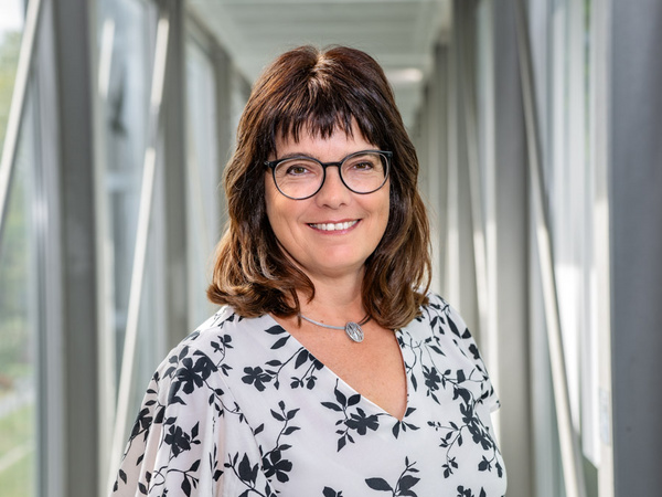 Eine Frau mit langen, braunen Haaren und Brille lächelt freundlich in die Kamera. Sie trägt ein blumiges Oberteil und steht in einem hellen, modernen Raum mit großen Fenstern im Hintergrund. (Text KI generiert)