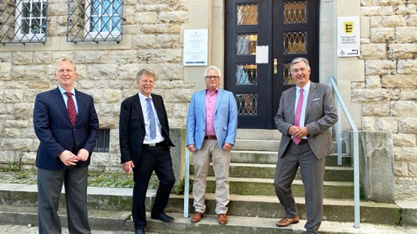 Quatre hommes en costume se tiennent devant l'entrée d'un bâtiment en pierre nommé "KLOBERAMT". Ils sourient, affichant une atmosphère amicale. L'un porte une chemise rose, un autre un costume clair. Des marches mènent à une porte en bois ornée de vitraux.