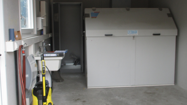 A spacious garage with a concrete floor features a large storage unit in the middle, a cleaning device on the left, and tools hung on the wall. A doorway leads to another room, and overhead lights illuminate the area.
