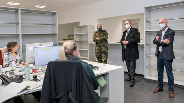 In an office setting, a woman sits at a desk with a computer, engaged in a discussion. Two men, one in military attire and the other in a suit, observe the conversation while wearing masks. A third man stands nearby, also in a suit, listening attentively.