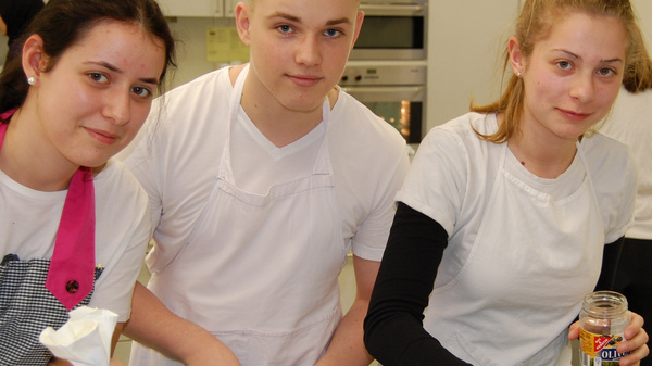 Three young people wearing white shirts and aprons work in a kitchen, preparing desserts. One holds a piping bag, while another adds ingredients from a jar to a tray of desserts. The setting appears busy with other individuals in the background.