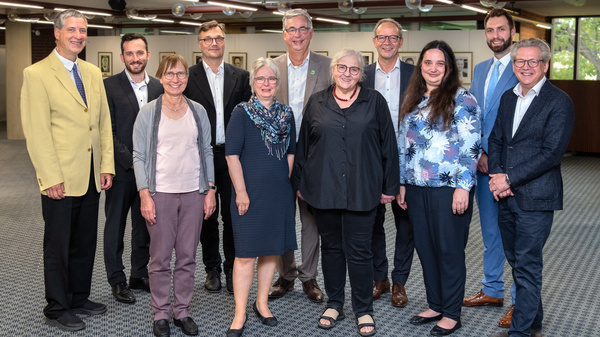 Eine Gruppe von zwölf Personen steht in einem Raum, lächelt und posiert für ein Foto. Die Personen sind unterschiedlich gekleidet und scheinen an einem offiziellen Anlass teilzunehmen. Im Hintergrund sind Bilder an den Wänden zu sehen.