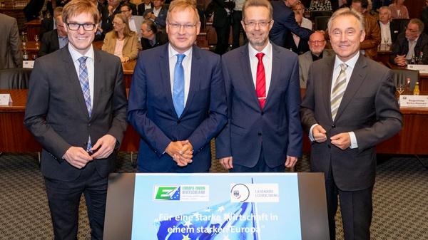 Four men in suits pose together in front of a presentation screen displaying the title "FüR eine starke Wirtschaft in einem starken Europa." The setting appears to be a formal event or conference, with an audience visible in the background.