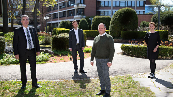 Four individuals stand in a park setting, surrounded by greenery and shrubs. They are dressed in formal and semi-formal attire, appearing engaged in conversation. The scene is bright and sunny, reflecting a professional yet relaxed atmosphere.