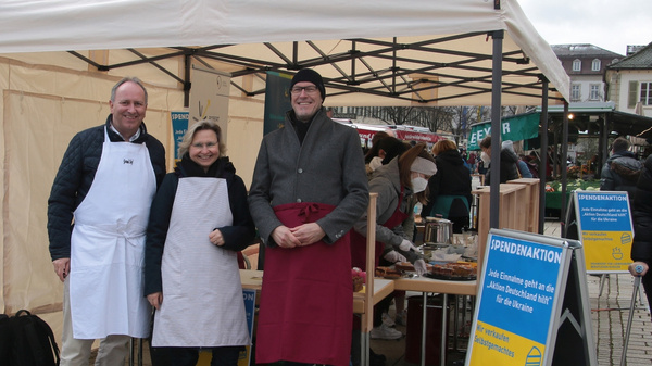 Drei Personen stehen lächelnd unter einem Marktstand mit einem weißen Zelt. Sie tragen Schürzen und präsentieren ihre Produkte. Im Hintergrund sind weitere Marktstände und eine belebte Platzatmosphäre sichtbar. Ein Informationsschild steht neben dem Stand.