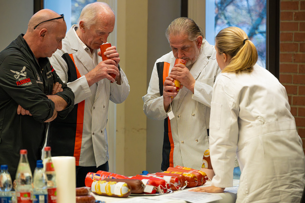 Vier Personen stehen um einen Tisch, auf dem verschiedene Wurstwaren liegen. Zwei Männer und eine Frau halten Verpackungen und betrachten sie aufmerksam. Eine Person sieht nachdenklich aus, während der andere einen Snack probiert. Der Fokus liegt auf der kulinarischen Analyse und dem Austausch von Erfahrungen.