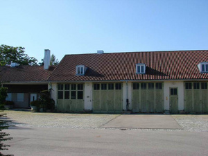Das Bild zeigt ein zweigeschossiges Gebäude mit einem roten Satteldach und mehreren Fenstern. Die Fassade ist teilweise mit Holzlatten vertäfelt. Vor dem Gebäude befindet sich eine asphaltierte Fläche. Die Umgebung ist mit Bäumen und strauchartiger Vegetation bewachsen. Der Himmel ist blau und klar.