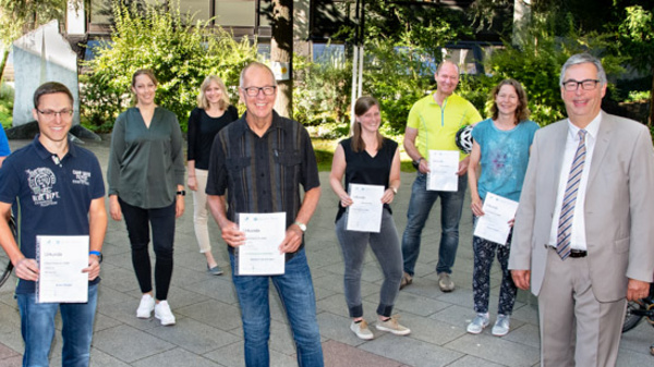 A group of eight individuals stands in a courtyard, each holding a certificate. They are accompanied by bicycles and a man in a suit. The scene suggests a celebration of an achievement or recognition event related to cycling or environmental awareness.
