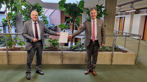 Deux hommes en costumes se tiennent debout dans un espace intérieur lumineux, entourés de plantes. L’un d’eux tend un document à l’autre, marquant un moment officiel. L’ambiance est professionnelle et heureuse.