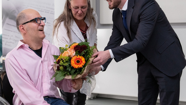 Ein Mann im Rollstuhl erhält einen Blumenstrauß von einem Mann im Anzug. Eine Frau mit langen Haaren beobachtet die Szene. Im Hintergrund ist ein Plakat sichtbar, das eine Botschaft über Geschichte enthält.