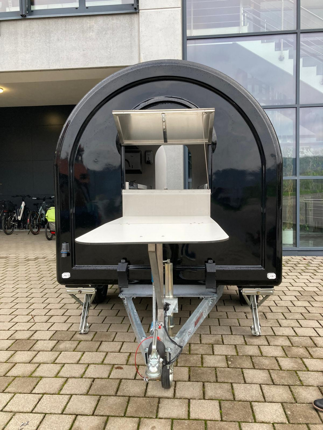 Ein kompakter, schwarzer Verkaufsanhänger mit einem aufklappbaren Thekentisch. Die Vorderseite enthält eine offene Fläche, die als Verkaufsfläche dient. Der Anhänger steht auf einem gepflasterten Boden vor einem Gebäude.