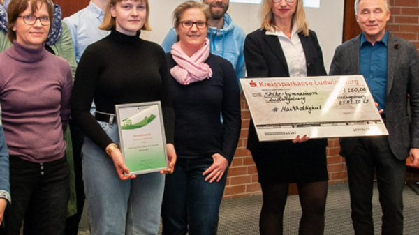 A group of people stands together in a room, holding a certificate and a large check. They appear to be celebrating a recognition or award. The background includes a presentation screen with the hashtag "#MachHalt," indicating a theme or campaign related to the event.