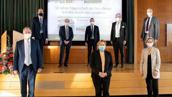 A group of seven people, wearing business attire and masks, stands on a stage in front of a screen displaying commemorative text for the 50th anniversary of vocational schools. The setting suggests a formal event or celebration.