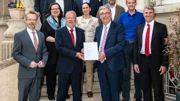 Eine Gruppe von 15 Personen steht auf einer Treppe vor einem Gebäude. Zwei Männer im Vordergrund halten ein Dokument in den Händen. Die Anwesenden sind formell gekleidet und lächeln. Im Hintergrund sind Fenster und Pflanzen sichtbar.