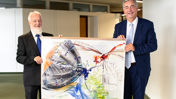 Deux hommes se tiennent debout, souriant, en tenant une grande peinture représentant des insectes colorés. L'un est barbu, vêtu d'un costume noir, l'autre porte un costume bleu et une cravate. L'arrière-plan est flou, suggérant un environnement de bureau.