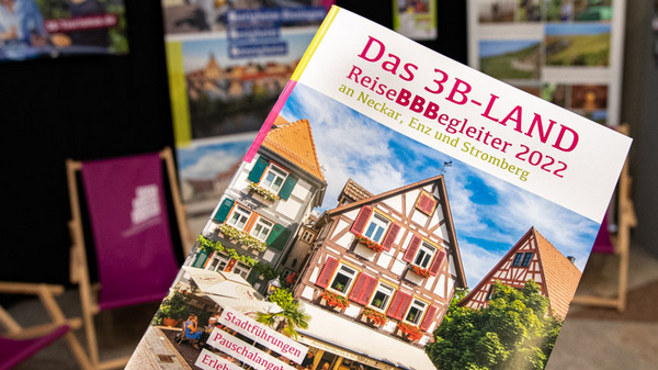 Ein Reiseführer für das 3B-Land, inklusive Bilder von malerischen Gebäuden. Der Titel „ReiseBBegleiter 2022“ ist prominent auf dem Cover abgebildet. Im Hintergrund sind Informationsstände mit weiteren Materialien und Plakaten zur Region sichtbar.