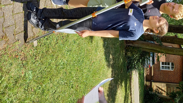 Deux personnes, un homme et une femme, utilisent un appareil de nivellement sur un terrain gazonné. Ils semblent concentrés sur leur tâche. L'environnement est boisé avec des arbres en arrière-plan.
