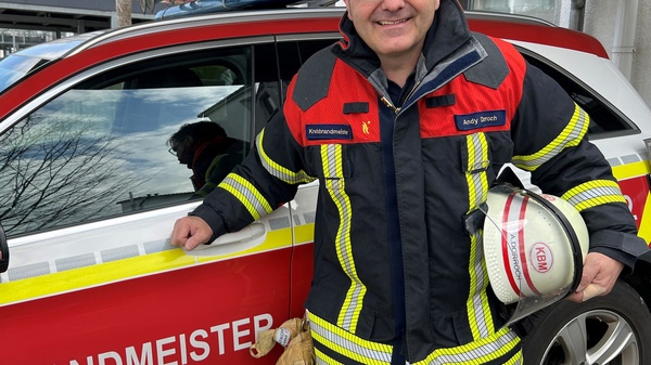 Ein Feuerwehrmann in uniform steht neben einem roten und gelben Einsatzfahrzeug, das die Aufschrift „KREISBRANDMEISTER“ trägt. Er hält einen Helm in der Hand und lächelt in die Kamera. Im Hintergrund sind Bäume und ein Gebäude sichtbar.