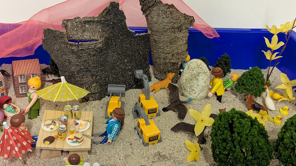 Eine blaue Kiste zeigt eine Spielszene mit Miniaturfiguren, die ein Picknick im Freien genießen. Es gibt einen Tisch mit Essen, Spielzeugautos, Bäume und Blumen. Im Hintergrund sind zerfallene Mauern zu sehen, die eine natürliche Umgebung schaffen.