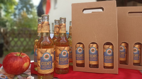 The image features several bottles of a beverage labeled "GIDRE," displayed next to a bright red apple and a cardboard six-pack carrier. The setting suggests a casual outdoor environment, highlighting the drink product.