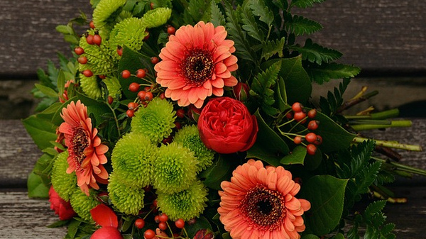 Un bouquet di fiori colorati composto da gerbere arancioni, rose rosse, fiori verdi e foglie verdi. Il mazzo è posato su una superficie di legno.