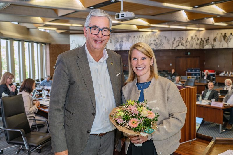 Zwei Personen stehen in einem Konferenzraum. Der Mann trägt ein graues Jackett und lächelt, während er eine Frau mit einem Blumenstrauß in der Hand anlächelt. Im Hintergrund sind weitere Personen und Tische zu sehen. (Text KI generiert)