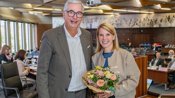 Zwei Personen stehen in einem Konferenzraum. Der Mann trägt ein graues Jackett und lächelt, während er eine Frau mit einem Blumenstrauß in der Hand anlächelt. Im Hintergrund sind weitere Personen und Tische zu sehen. (Text KI generiert)
