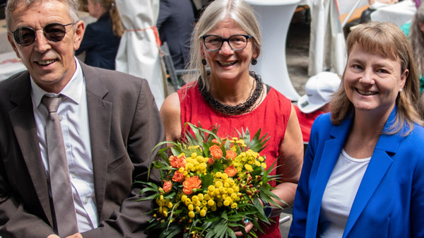 Three individuals pose together for a photo outdoors. The first person, wearing sunglasses and a suit, smiles on the left. In the center, a woman holds a colorful bouquet and wears a red top. The third person, dressed in a blue blazer, stands on the right. People mingle in the background.