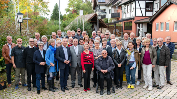 A large group of approximately 40 people stands together in a courtyard surrounded by buildings and greenery. The individuals are dressed casually, with some wearing coats and jackets. A mix of genders and ages is represented, and they appear to be smiling and enjoying the moment.