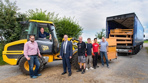 Un groupe de sept personnes se tient près d'une grue jaune JCB et d'un camion de livraison. Certains sourient, tandis qu'un homme conduit la grue. En arrière-plan, des arbres verdoyants et un chargement en bois dans le camion. L'ambiance est conviviale et professionnelle.