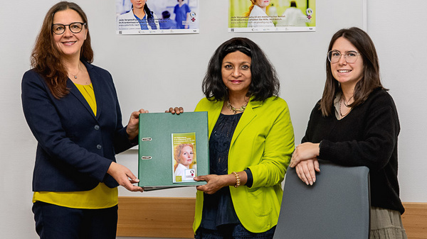 Trois femmes se tiennent debout dans un bureau, chacune portant des vêtements aux teintes de vert. Elles tiennent un dossier avec une image d'une femme au centre. Des affiches en arrière-plan traitent de sujets médicaux. L'atmosphère est collaborative et engageante.