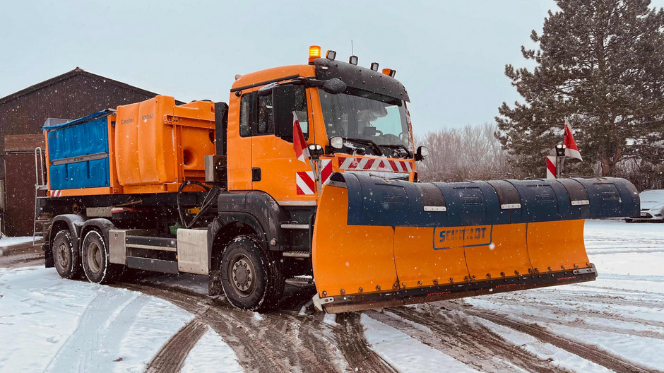 A snowplow truck is parked on a snowy road. The vehicle is orange with a front-mounted blade for clearing snow. It has a blue container in the back, likely for holding salt or sand. Surrounding trees and a building are partially visible in the background.