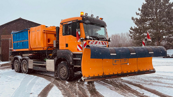 A snowplow truck is parked on a snowy road. The vehicle is orange with a front-mounted blade for clearing snow. It has a blue container in the back, likely for holding salt or sand. Surrounding trees and a building are partially visible in the background.