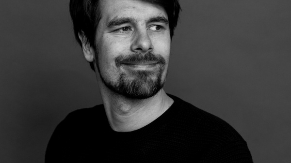 Ritratto di un uomo con barba scura e capelli lunghi, che sorride serenamente, indossando un maglione nero. Lo sfondo è neutro e scuro, creando un contrasto con la sua espressione rilassata e amichevole.