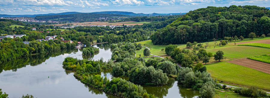 Header Bild: Eine weite Landschaft mit einem ruhigen Fluss, der von üppigem Grün gesäumt ist. Auf der anderen Seite sind sanfte Hügel und Felder zu sehen, während der Himmel mit einigen Wolken bedeckt ist. Die Szene strahlt eine friedliche, natürliche Umgebung aus.