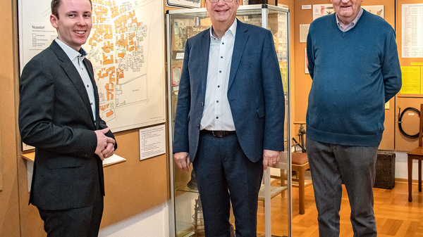 Drei Männer stehen in einem Museum mit historischen Ausstellungen zu „Alte Nussdorf“. Sie lächeln und interagieren, während im Hintergrund Informationen und Fotos an Wänden präsentiert werden. Die Atmosphäre ist informativ und einladend, mit Fokus auf lokale Geschichte.