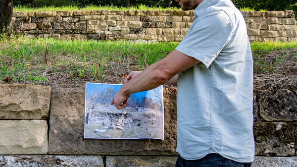 Ein Mann steht an einer Steinmauer und hält ein Bild hoch, das eine alte, zerstörte Landschaft zeigt. Er zeigt mit dem Finger auf einen Bereich des Bildes, während im Hintergrund eine grüne Umgebung mit Bäumen zu sehen ist.