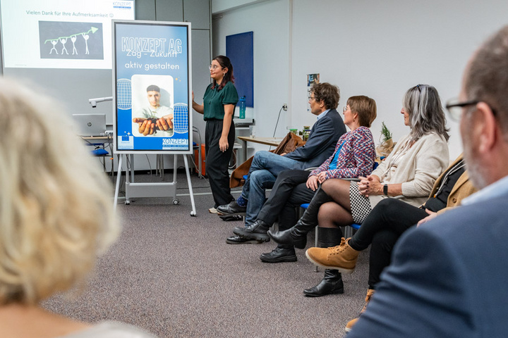 Eine Präsentation in einem Seminarraum, in dem eine Frau vor einer Gruppe von sechs Personen spricht. Im Hintergrund ist ein Plakat mit dem Text „KONZEPT.AG aktiv gestalten“ zu sehen, während die Zuhörer aufmerksam zuhören.