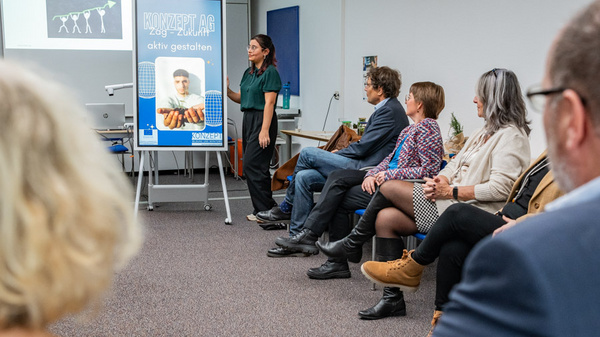 Eine Präsentation in einem Seminarraum, in dem eine Frau vor einer Gruppe von sechs Personen spricht. Im Hintergrund ist ein Plakat mit dem Text „KONZEPT.AG aktiv gestalten“ zu sehen, während die Zuhörer aufmerksam zuhören.