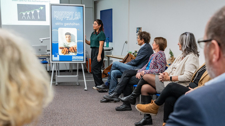 Eine Präsentation in einem Seminarraum, in dem eine Frau vor einer Gruppe von sechs Personen spricht. Im Hintergrund ist ein Plakat mit dem Text „KONZEPT.AG aktiv gestalten“ zu sehen, während die Zuhörer aufmerksam zuhören.